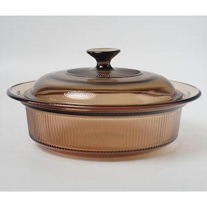 May include: A round, amber-coloured glass casserole dish with a matching lid. The dish features vertical ribbed detailing along the exterior sides, while the lid has a central knob handle and a radial sunburst pattern on the top surface.