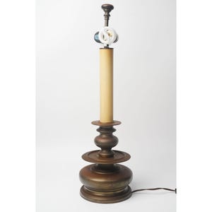 May include: A bronze candlestick lamp with a beige cylindrical shade and a decorative base. The lamp has a bulb and a power cord. The base features a tiered design with circular platforms and rounded sections.