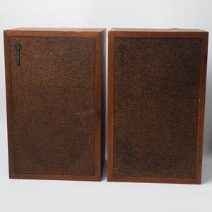 May include: A pair of rectangular wooden speakers with dark brown textured fabric grilles. The left speaker features a small black circular logo emblem in the upper left corner. Both units have a smooth, medium-toned wood frame surrounding the front.
