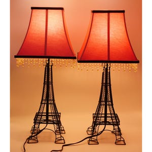 Peut inclure: Deux lampes de table décoratives avec des abat-jours rouges et des bases en forme de Tour Eiffel noires. Les abat-jours carrés sont bordés d'une bordure foncée et de perles de cristal. Les lampes sont reliées par un câble noir.