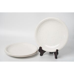 May include: A set of three round ceramic plates with a speckled off-white finish. One plate is displayed upright on a dark brown wooden stand, while two identical plates are stacked horizontally on the surface next to the upright plate.