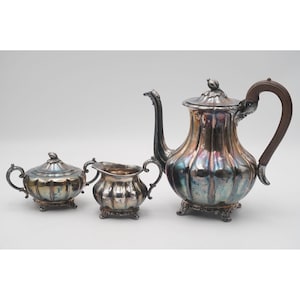 Op de afbeelding: Een zilveren theeservies met een theepot, melkkan en suikerpot. De theepot heeft een houten handvat en een decoratief deksel. De set heeft een verouderde patina, met iriserende kleuren op het oppervlak. De melkkan en suikerpot hebben een bijpassend ontwerp.
