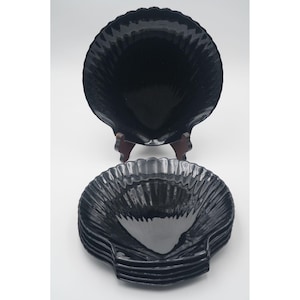 Könnte beinhalten: Ein Set schwarzer, muschelförmiger Servierschalen. Eine große Schale wird auf einem Holzständer präsentiert, darunter ein Stapel kleinerer Schalen. Die Schalen haben eine glänzende Oberfläche und eine geriffelte Textur.