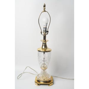 May include: A table lamp featuring a clear cut glass body with a diamond pattern, a polished gold-tone metal base, and a matching gold-tone metal neck. The lamp includes a harp, a finial at the top, and a white light bulb installed in the socket.