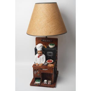 Könnte beinhalten: Eine originelle Lampe mit einer Kochfigur in einer Küchenszene. Die Lampe hat einen beigefarbenen Lampenschirm aus Jute und einen Holzsockel mit einem Koch, einem Ofen und Pizzazutaten. Das Wort "PIZZA" steht über dem Koch. Die Lampe ist ein dekorativer Artikel.