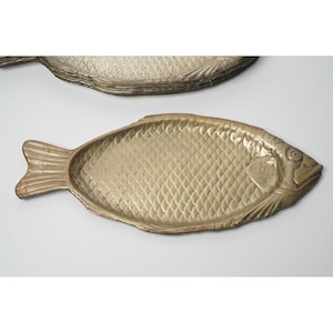 Puede incluir: Dos bandejas decorativas con forma de pez. Las bandejas están hechas de un material metálico con acabado dorado. Las escamas de los peces están detalladas, y las bandejas tienen cola y cabeza. Las bandejas están diseñadas para servir alimentos.