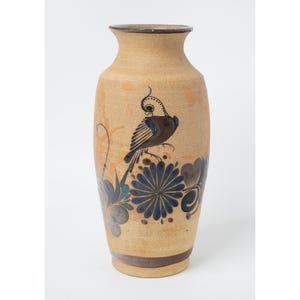 May include: A tall, cylindrical ceramic vase with a light tan, speckled finish. The vase features hand-painted dark blue and brown floral motifs and a central illustration of a crested bird perched above a large, detailed flower blossom.