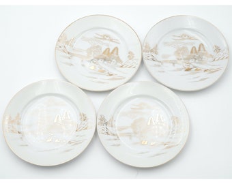May include: Set of four white ceramic plates with gold trim and a Japanese-inspired design. Each plate features a landscape scene with a bridge, trees, and buildings.