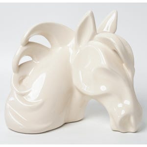 May include: A glossy, off-white ceramic sculpture depicting the head and neck of a horse. The piece features smooth, stylized curves that define the mane, ears, and facial structure, creating a continuous, fluid form with a reflective surface finish.