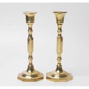 May include: Two brass candlesticks with a hexagonal base and a flared top. The candlesticks have a classic design with a slender stem and a polished finish. The candlesticks are approximately 10 inches tall.
