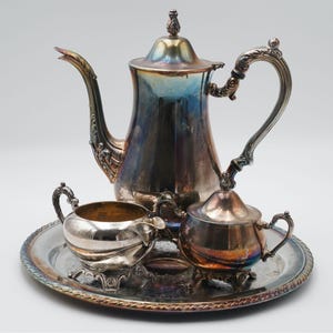 Op de afbeelding: Een zilveren theeservies bestaande uit een theepot, melkkan en suikerpot, gepresenteerd op een bijpassend dienblad. De set heeft sierlijke handvatten en een decoratief ontwerp. De theepot heeft een hoge, gebogen tuit.