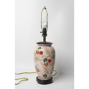 Può includere: Una lampada da tavolo in ceramica con base color crema caratterizzata da un motivo dipinto di foglie rosse, verdi e dorate. La lampada presenta una base circolare e un bordo superiore marrone scuro, un'arpa in metallo e una lampadina bianca.