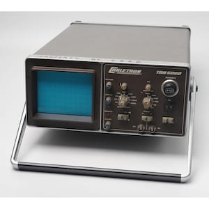 Puede incluir: Un dispositivo electrónico vintage gris y negro, un Coletron TDR 5000, con una pantalla de cuadrícula azul y numerosos diales y perillas. El dispositivo tiene un asa plateada y el texto "PROPERTY OF C S S C" es visible.