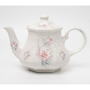 Puede incluir: Tetera de cerámica blanca con un patrón de celosía y un diseño floral rosa. La tetera tiene un pico curvo, un asa redondeada y una tapa con un pequeño pomo. El diseño floral incluye peonías rosas y capullos con hojas verdes y grises.