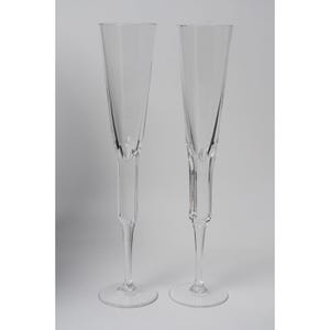 May include: A pair of clear glass champagne flutes featuring tall, slender bowls that taper toward the base. Each flute has a distinct faceted stem design that transitions into a smooth, circular foot, providing a stable base for these elegant vessels.