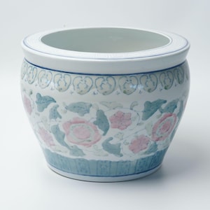 Puede incluir: Una maceta redonda de cerámica blanca con una amplia abertura. La maceta presenta un diseño floral con flores rosas y hojas azul-verdosas. Bandas azules con patrones decorativos rodean la parte superior e inferior.
