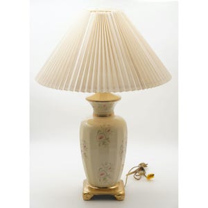 May include: A vintage table lamp with a cream-colored pleated lampshade. The base is a cream-colored ceramic vase with pink floral accents and gold trim. The lamp sits on a gold square base. The lamp has a cord with a yellow plug.