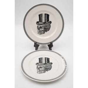 May include: Two white ceramic plates with a black and gray skull wearing a top hat design. The plates have a gray border with a decorative pattern. One plate is displayed on a wooden stand.