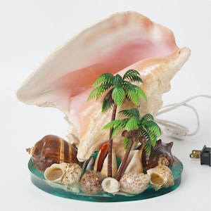 May include: A decorative lamp featuring a large, cream and pink conch shell as the primary shade. The base is a circular, translucent green resin platform adorned with smaller brown and white seashells and two miniature green plastic palm trees.