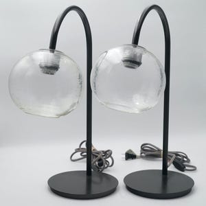 May include: Two modern table lamps. Each lamp features a clear, spherical glass shade and a black, curved metal arm. The lamps sit on round, black bases and have gray cords with plugs.