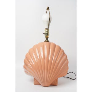 May include: A table lamp featuring a glossy, peach-colored ceramic base shaped like a large scallop shell. The lamp includes a brass-toned metal socket assembly with a white light bulb attached to the top, and a black electrical cord extending.