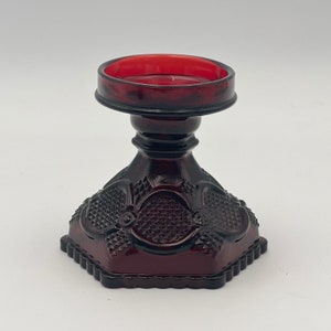 May include: A red glass candle holder with a scalloped edge and a decorative design. The holder has a round top and a wide base.