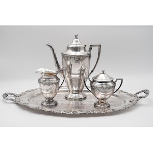 Puede incluir: Un juego de té de plata sobre una bandeja decorativa. El juego incluye una tetera, una crema y un azucarero. La bandeja y las piezas tienen detalles ornamentados. El juego se muestra sobre un fondo blanco.
