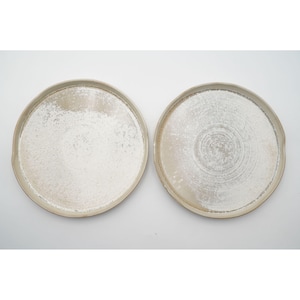 May include: Two round ceramic plates with a speckled, off-white glaze. The plates have a slightly raised rim and a textured surface, creating a rustic aesthetic. The plates are set against a plain white background.