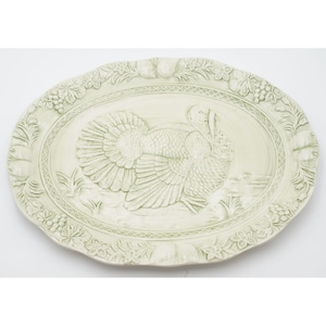 Puede incluir: Plato ovalado de cerámica blanquecina con un diseño en relieve de un pavo. El plato tiene un borde festoneado con motivos florales y frutales. El pavo está centrado, con texturas detalladas de plumas y cuerpo, ideal para servir una comida festiva.