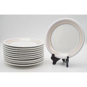 May include: A stack of white ceramic plates with a colorful striped border. Each plate has a design of red, yellow, green, and blue stripes. One plate is displayed on a black stand. The plates are suitable for everyday use.