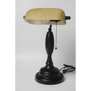 May include: A black banker's desk lamp with a beige, curved shade. The lamp has a round base and a slender, curved stem. A pull chain hangs from the shade. The lamp is plugged into an electrical outlet.