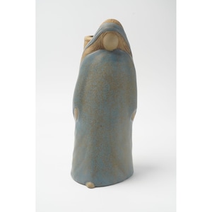May include: A ceramic sculpture of a robed figure in a light blue hue, with a hood and long, flowing hair. The figure has a serene expression and is holding a small object. The sculpture has a textured surface and is set against a white background.