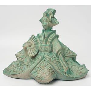 May include: A teal ceramic figurine of a seated geisha. The figure wears an elaborate headdress and kimono with intricate patterns. She holds a fan in her left hand. The sculpture has a weathered, aged appearance.