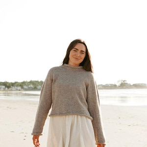 Puede incluir: Un suéter beige de manga larga con cuello redondo y dobladillo sin rematar, combinado con una minifalda blanca plisada y botas de goma verde oscuro hasta la rodilla. El conjunto se lleva en una playa de arena bajo un cielo soleado.