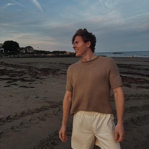 Puede incluir: Una persona con una camisa de punto marrón de manga corta y pantalones cortos color crema en una playa de arena. El cielo es azul con nubes tenues. La persona sonríe, lo que sugiere un estilo relajado e informal. La escena es junto al mar.