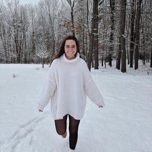 May include: A person wearing a white oversized turtleneck sweater, black tights, and tan lace-up boots, walking in the snow. The background features a snowy forest with bare trees.