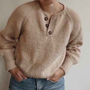 May include: A beige knitted sweater with a v-neck and a button closure. The sweater has long sleeves and a relaxed fit.