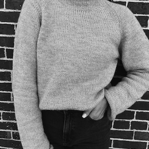May include: A light grey knitted jumper with a high, ribbed collar and long sleeves. The jumper has a relaxed fit and is made of a textured yarn. The sleeves have ribbed cuffs. The person is wearing black jeans.