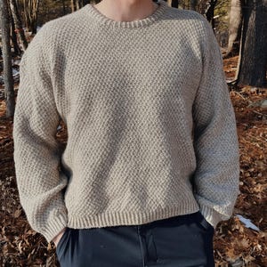 Könnte beinhalten: Ein beiger Strickpullover mit einem strukturierten Muster. Der Pullover hat einen Rundhalsausschnitt und lange Ärmel.
