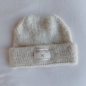 Pode incluir: Um gorro de malha branco com uma etiqueta que diz "we are knitters™ 100% natural fibers" e "Proudly handmade by myself".