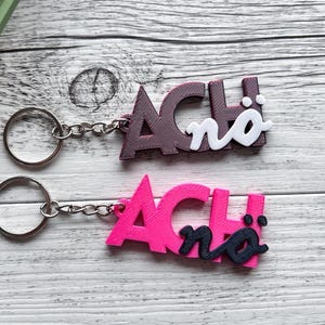 Könnte beinhalten: Zwei Schlüsselanhänger mit dem Wort "ACH!" in einer stilisierten Schriftart. Einer ist magenta mit schwarzen Akzenten, der andere ist dunkelbraun mit weißen Akzenten. Beide haben silberne Schlüsselringe und Ketten.