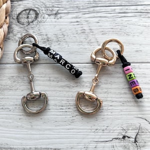 Könnte beinhalten: Zwei Schlüsselanhänger mit Trensen-Designs. Einer ist silberfarben mit dem Namen "MARCO" in schwarz-weißen Perlen. Der andere ist goldfarben mit dem Namen "INES" in bunten Perlen. Beide haben eine schwarze Kordel.