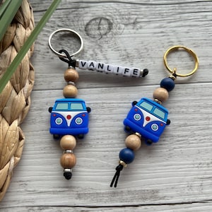 Könnte beinhalten: Zwei blaue und weiße Van-Schlüsselanhänger mit Holzperlen und einem schwarzen Quasten. Ein Schlüsselanhänger hat das Wort "VANLIFE" in weißen Buchstaben.