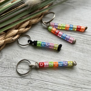 May include: Keychains with rainbow-colored beads spelling out names. The names include "FELIX", "DANIELA", "FRANZISKA", and "STELZER". Each keychain has a silver ring and a cord or string attachment.