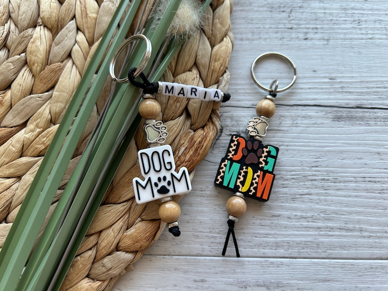 May include: Two keychains with wooden beads and a dog paw charm. One keychain has a white charm with the text "DOG MOM" and the other has a colourful charm with the text "DOG MOM".