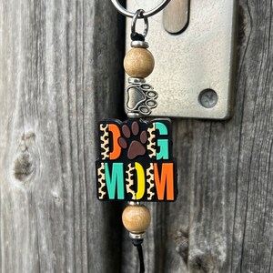 May include: A colourful keychain with the words "DOG MOM" in a black, orange, yellow and teal colour scheme. The keychain has a silver paw print charm and wooden beads.