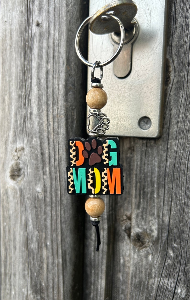 May include: A colourful keychain with the words "DOG MOM" in a black, orange, yellow and teal colour scheme. The keychain has a silver paw print charm and wooden beads.