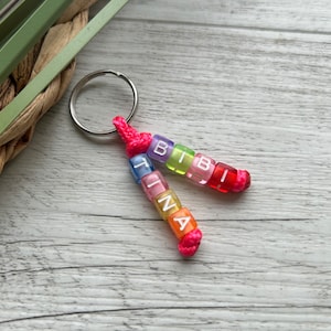 May include: A personalized keychain featuring two strands of colorful beads spelling out "TINA" and "BIBI". The beads are in various colors, including pink, orange, yellow, green, and blue, with white letters. The keychain is attached to a silver ring with a pink cord.