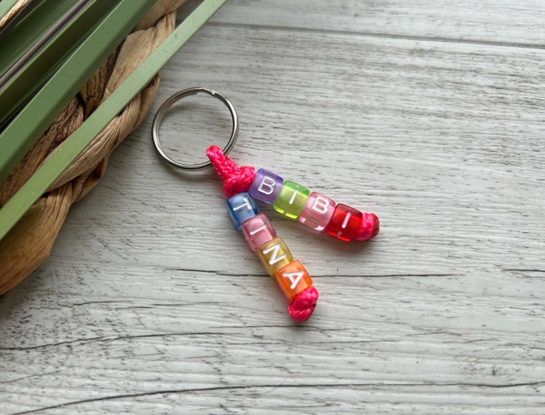 May include: A personalized keychain featuring two strands of colorful beads spelling out "TINA" and "BIBI". The beads are in various colors, including pink, orange, yellow, green, and blue, with white letters. The keychain is attached to a silver ring with a pink cord.