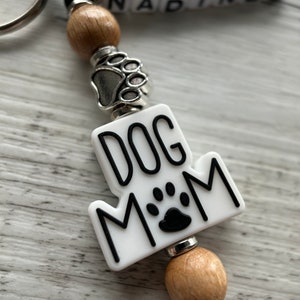 May include: White keychain with black text that reads "Dog Mom" and a black paw print. The keychain has a silver paw print charm and wooden beads.